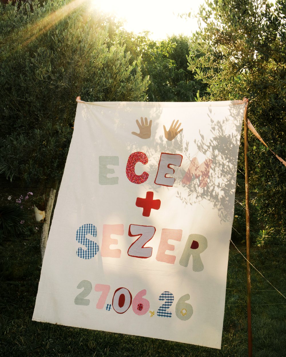 Bahçede gerilmiş bez banner: kumaş harflerle iki isim, aralarında kırmızı artı, altında tarih